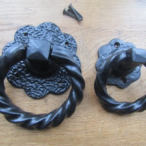 May include: Two black door knockers with twisted ring handles and decorative backplates. The knockers are made of metal and are shown with a few screws, ready for installation. The backplates have a floral design.