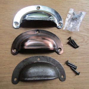 May include: Three metal cup pull handles in silver, copper, and dark grey finishes, with mounting screws. The handles are arranged on a light brown wooden surface, showcasing their different colours and textures. These are cabinet hardware.