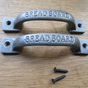 Puede incluir: Dos tiradores de armario de metal gris con las palabras "Bread Board" en relieve. Los tiradores están sobre una superficie de madera con dos tornillos.