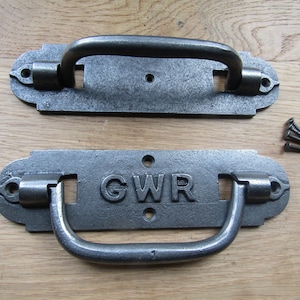 以下が含まれることがあります： GWR と刻印された鋳鉄製のドアハンドル2個。ハンドルは濃いグレーで、シンプルで長方形のデザインです。ハンドルは木製の表面に置かれています。