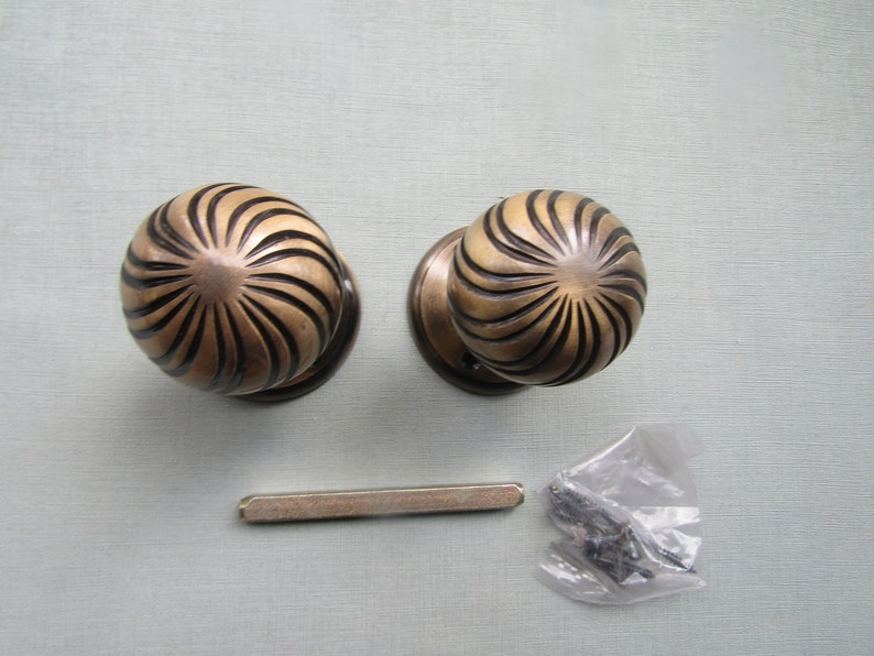 May include: Two antique brass door knobs with a sunburst design. The knobs are round and have a textured surface. The knobs are accompanied by a metal spindle and screws.
