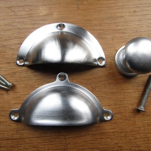 Puede incluir: Tres pomos de armario plateados, dos son en forma de taza y uno es redondo. Los pomos en forma de taza tienen una superficie lisa y redondeada. El pomo redondo tiene una superficie lisa y redondeada. Los pomos están sobre una superficie de madera.