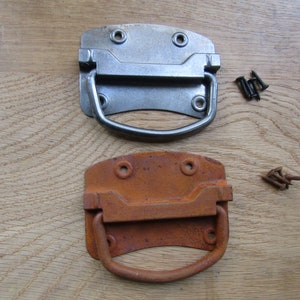May include: Two metal drawer pulls, one silver and one rusty orange, with a rounded rectangular shape and a D-shaped handle. Both pulls are shown on a wooden surface with screws.
