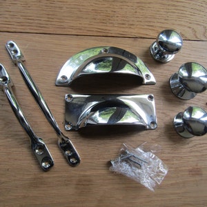 Può includere: Un set di ferramenta per armadi in cromo, tra cui due maniglie, due maniglie a coppa e due pomelli. La ferramenta è disposta su una superficie in legno.