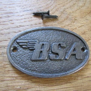 May include: A grey cast iron oval sign with the word "BSA" and a winged logo. The sign has two screw holes and comes with two screws.