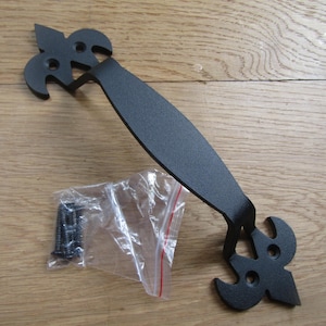 May include: Black metal door handle with a fleur-de-lis design. The handle is attached to a wooden surface and comes with screws.