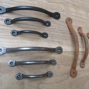 Op de afbeelding: Assortiment van zwarte en roestige metalen ladegrepen. De grepen zijn gebogen en hebben afgeronde uiteinden.