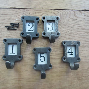 May include: Five black metal hooks with a square frame and a number from 1 to 5 in a white rectangle inside the frame. The hooks are on a wooden surface.