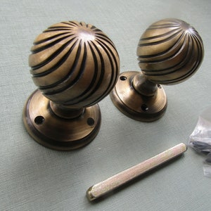 May include: A pair of antique brass door knobs with a spiral design. The knobs are attached to round brass plates. The image also shows a metal spindle and screws.
