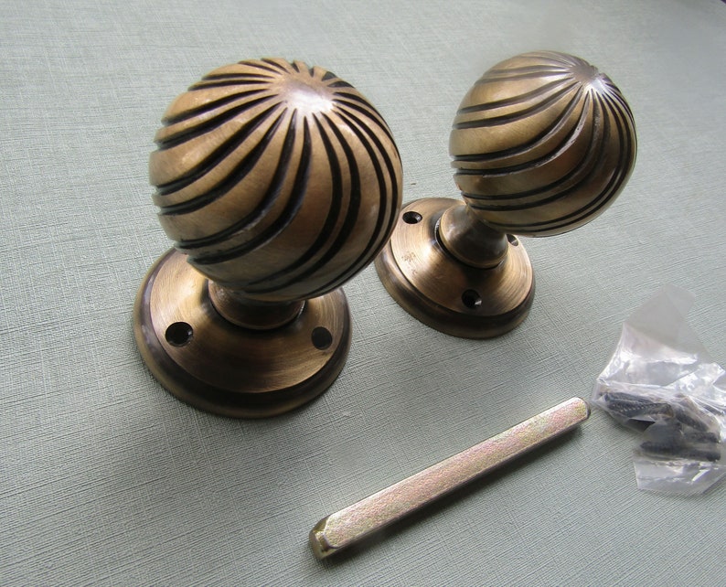 May include: A pair of antique brass door knobs with a spiral design. The knobs are attached to round brass plates. The image also shows a metal spindle and screws.