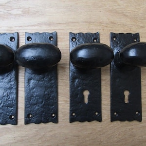 Peut inclure: Quatre poignées de porte noires avec des poignées ovales et des trous de serrure. Les poignées sont montées sur des plaques rectangulaires avec un bord décoratif. Les plaques ont chacune deux trous de vis. Les poignées sont présentées sur une surface en bois avec des vis et une tige métallique.