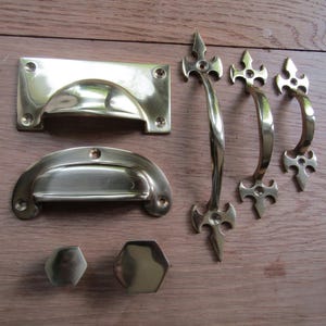 Könnte beinhalten: Eine Sammlung von Messingbeschlägen, darunter Schubladengriffe, Griffe und Knöpfe. Das Set besteht aus einem rechteckigen Griff, einem halbmondförmigen Griff, zwei sechseckigen Knöpfen und drei verzierten Griffen mit dekorativen Zierknöpfen.