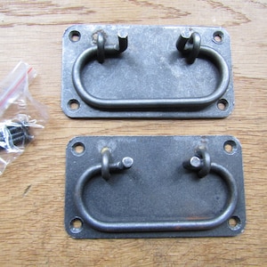 Könnte beinhalten: Zwei schwarze Metall-Schubladengriffe mit rechteckiger Basis und abgerundetem Schleifen-Griff. Die Griffe sind auf einer Holzoberfläche mit Schrauben dargestellt.