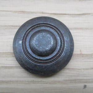 May include: A round, dark gray metal knob with concentric circles. The knob has a slightly weathered appearance, suggesting an antique or vintage style. The hardware is set against a light wood grain background.