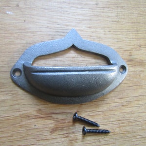 May include: A cast iron drawer pull with a curved handle and decorative top, shown with two black screws. The hardware has a dark grey finish and is designed for furniture or cabinetry.