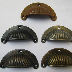 May include: A collection of five antique-style, half-moon drawer pulls in various finishes. The pulls are made of metal and feature a ribbed design. Finishes include rusty brown, black, gold, and bronze tones. Each pull has three screw holes.