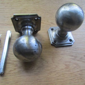 May include: Two silver door knobs with square backplates. The knobs are round and have a smooth, polished finish. The backplates have a decorative edge.