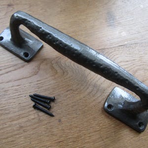 May include: A dark gray, hand-forged iron door pull with a textured finish. The handle is curved and attached to rectangular mounting plates with pre-drilled holes. Includes a set of black screws for installation. Ideal for rustic decor.