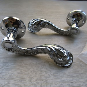 Może przedstawiać: Dwie chromowane klamki drzwiowe z ozdobnymi, dekoracyjnymi wzorami. Klamki są zakrzywione i mają okrągłą podstawę.