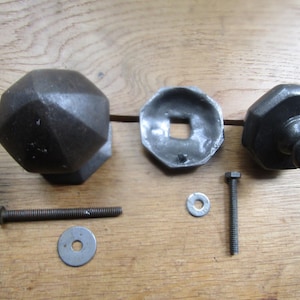May include: A selection of antique black metal cabinet hardware components, including two octagonal knobs, a square-holed backplate, screws, and washers. The hardware is arranged on a wooden surface, showing its aged appearance.