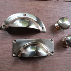 Pode incluir: Quatro peças de ferragens para armários de latão, incluindo duas puxadores de copo e duas maçanetas redondas. Os puxadores de copo são retangulares com uma borda arredondada e um centro curvo e embutido. As maçanetas são redondas com uma superfície lisa ligeiramente abobadada.