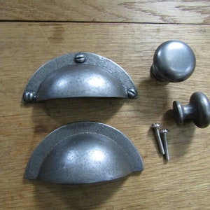 Może przedstawiać: Zestaw okuć do szafek w stylu antycznym, w tym dwa uchwyty do szuflad w kształcie półksiężyca, dwa okrągłe gałki i dwie śruby. Okucia mają zwietrzałe, metaliczne wykończenie, na drewnianej powierzchni.