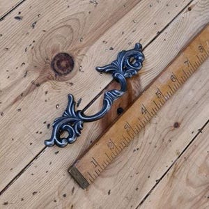 Może przedstawiać: Ozdobny, ciemny metalowy uchwyt do szuflady z zakrzywionym uchwytem i dekoracyjnymi zdobieniami. Uchwyt ma około 15 cm długości i spoczywa na drewnianej linijce i drewnianej powierzchni.