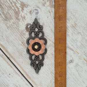 Może przedstawiać: Dekoracyjny uchwyt do szafki z ciemnoszarą, ozdobną metalową płytką tylną. Uchwyt ma brzoskwiniowy wzór kwiatowy z ciemnobrązowym środkiem i złotym środkiem. Obok uchwytu znajduje się drewniana linijka.