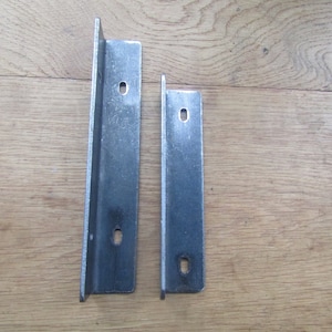 Może przedstawiać: Dwa metalowe wsporniki półki, jeden większy od drugiego, z otworami montażowymi. Wsporniki są srebrne i mają lekko fakturowaną powierzchnię.