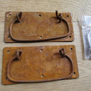 May include: Two rusty metal drawer pulls with a rounded rectangular shape and a loop handle. The pulls are on a wooden surface with a bag of screws.