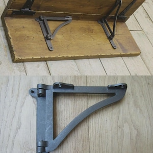 Pode incluir: Uma mesa dobrável de madeira com dobradiças e suportes de metal. A mesa é feita de madeira marrom clara e tem formato retangular. Os suportes de metal são cinza escuro e triangulares. A mesa está aberta.