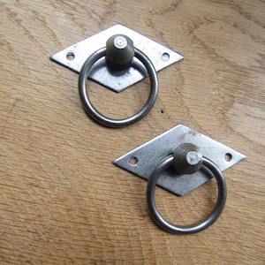 Pode incluir: Duas puxadores de gaveta de diamante prateados com alças de anel. As puxadores têm um aspeto ligeiramente envelhecido e rústico.