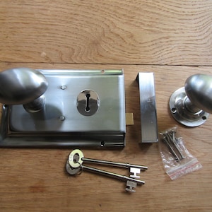 Puede incluir: Un juego de pomos de puerta de plata con dos pomos ovalados, una placa de cerradura rectangular, una placa de golpe, dos llaves y tornillos. El juego está sobre una superficie de madera.