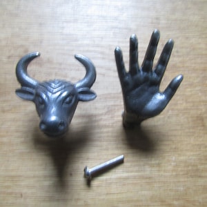 May include: Two metal cabinet knobs, one shaped like a bull's head and the other like a hand. The bull's head is black and the hand is gray. A silver screw is included.