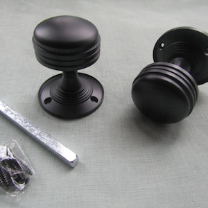 Puede incluir: Dos pomos de puerta negros con un diseño acanalado, una barra de metal y una pequeña bolsa de tornillos. Los pomos tienen una base redonda y una forma cilíndrica. Herrajes para puertas.