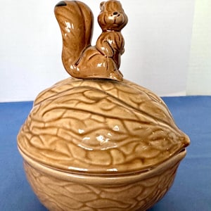 May include: A ceramic nut-shaped container with a brown finish. A small squirrel figurine sits atop the lid. The container has a detailed, textured surface, and the squirrel has a glossy finish. The overall design is whimsical and decorative.