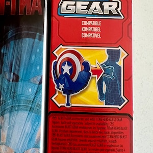 Pu&ograve; includere: Scatola rossa con la scritta "BLAST GEAR" e la parola "COMPATIBLE" in pi&ugrave; lingue. L'illustrazione presenta uno scudo e un contorno di figura, che suggerisce un accessorio giocattolo.