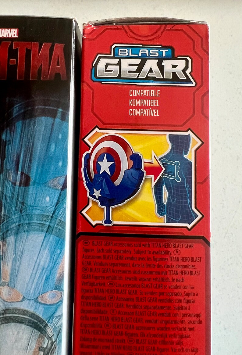 Pu&ograve; includere: Scatola rossa con la scritta "BLAST GEAR" e la parola "COMPATIBLE" in pi&ugrave; lingue. L'illustrazione presenta uno scudo e un contorno di figura, che suggerisce un accessorio giocattolo.