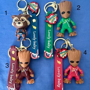 May include: Four keychains featuring characters from the Marvel Cinematic Universe. The keychains feature Rocket Raccoon, Groot, and Captain America. The keychains are made of plastic and have a metal ring for attaching to keys. The keychains are all different colors and have a "Lovely baby" tag.