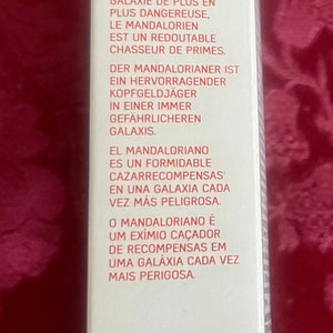 Puede incluir: Una caja rectangular blanca con el logo de Star Wars y el texto "The Mandalorian". La caja contiene texto en varios idiomas que describe a The Mandalorian como un cazarrecompensas en una galaxia peligrosa. El logo de Disney est&aacute; en la parte inferior.