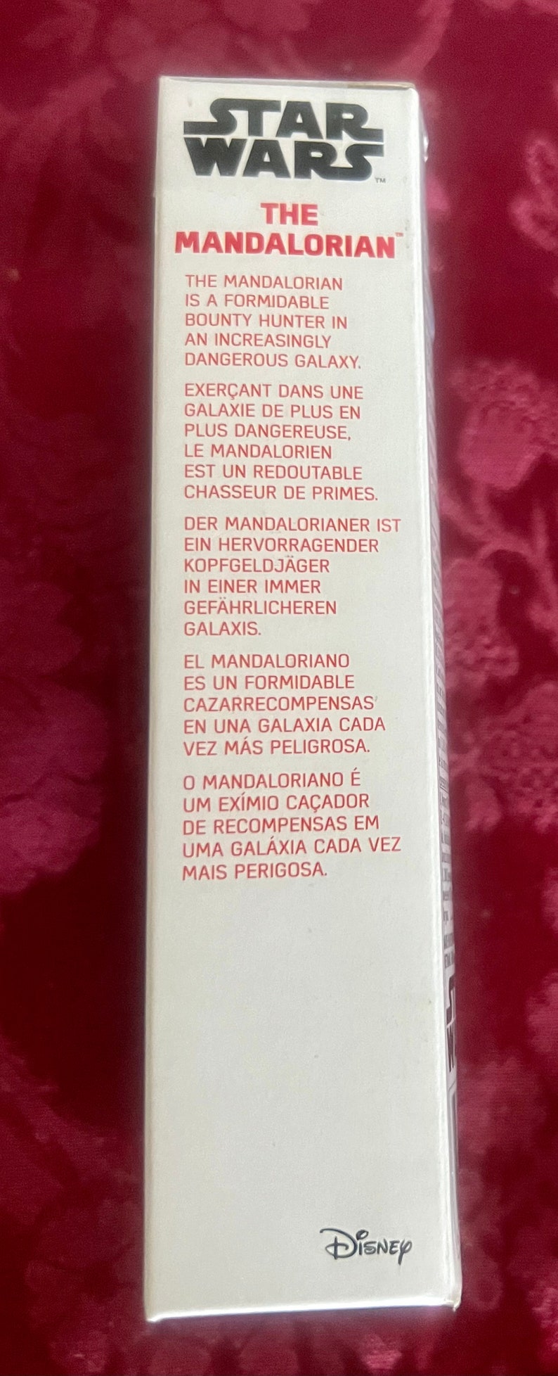 Puede incluir: Una caja rectangular blanca con el logo de Star Wars y el texto "The Mandalorian". La caja contiene texto en varios idiomas que describe a The Mandalorian como un cazarrecompensas en una galaxia peligrosa. El logo de Disney est&aacute; en la parte inferior.