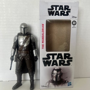 Puede incluir: Una figura de acci&oacute;n de Mandalorian con casco y armadura plateados, junto a su caja tem&aacute;tica de Star Wars. La caja presenta el logotipo de Star Wars y las palabras "The Mandalorian".