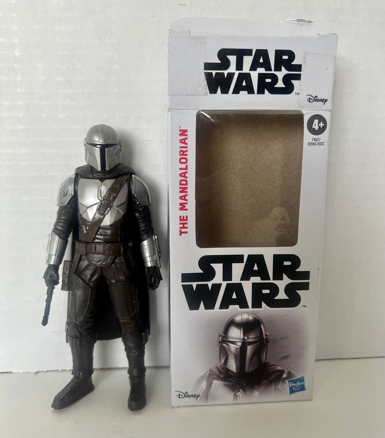 Puede incluir: Una figura de acci&oacute;n de Mandalorian con casco y armadura plateados, junto a su caja tem&aacute;tica de Star Wars. La caja presenta el logotipo de Star Wars y las palabras "The Mandalorian".