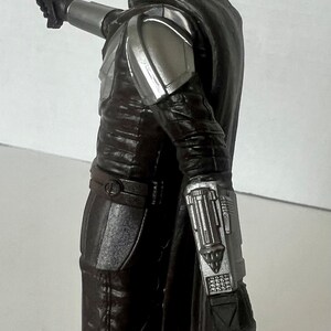 Puede incluir: Figura de acci&oacute;n detallada de un personaje de una serie de ciencia ficci&oacute;n. La figura viste una armadura marr&oacute;n oscuro con detalles plateados, un casco y una capa negra. Sostiene una pistola bl&aacute;ster.