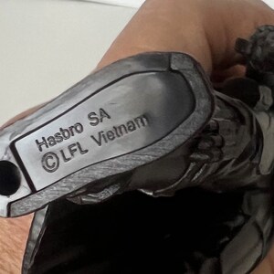 Puede incluir: Primer plano de una figura de acci&oacute;n gris oscuro con el texto "Hasbro SA &copy; LFL Vietnam" impreso en la parte inferior. La figura se sostiene en una mano.