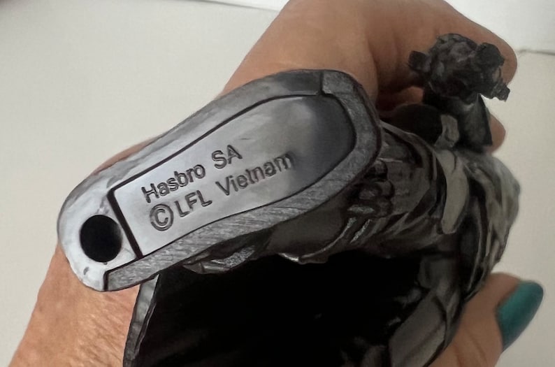 Puede incluir: Primer plano de una figura de acci&oacute;n gris oscuro con el texto "Hasbro SA &copy; LFL Vietnam" impreso en la parte inferior. La figura se sostiene en una mano.
