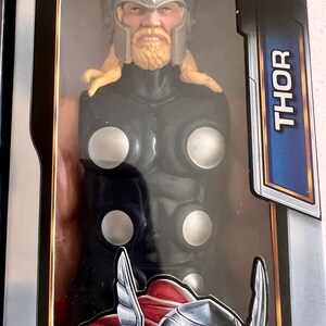 Pu&ograve; includere: Una action figure di Thor in una scatola. La figura ha un elmo argentato, capelli e barba biondi e un costume nero e argento. La scatola ha la scritta "Thor" in grandi lettere blu e il logo "Blast Gear".
