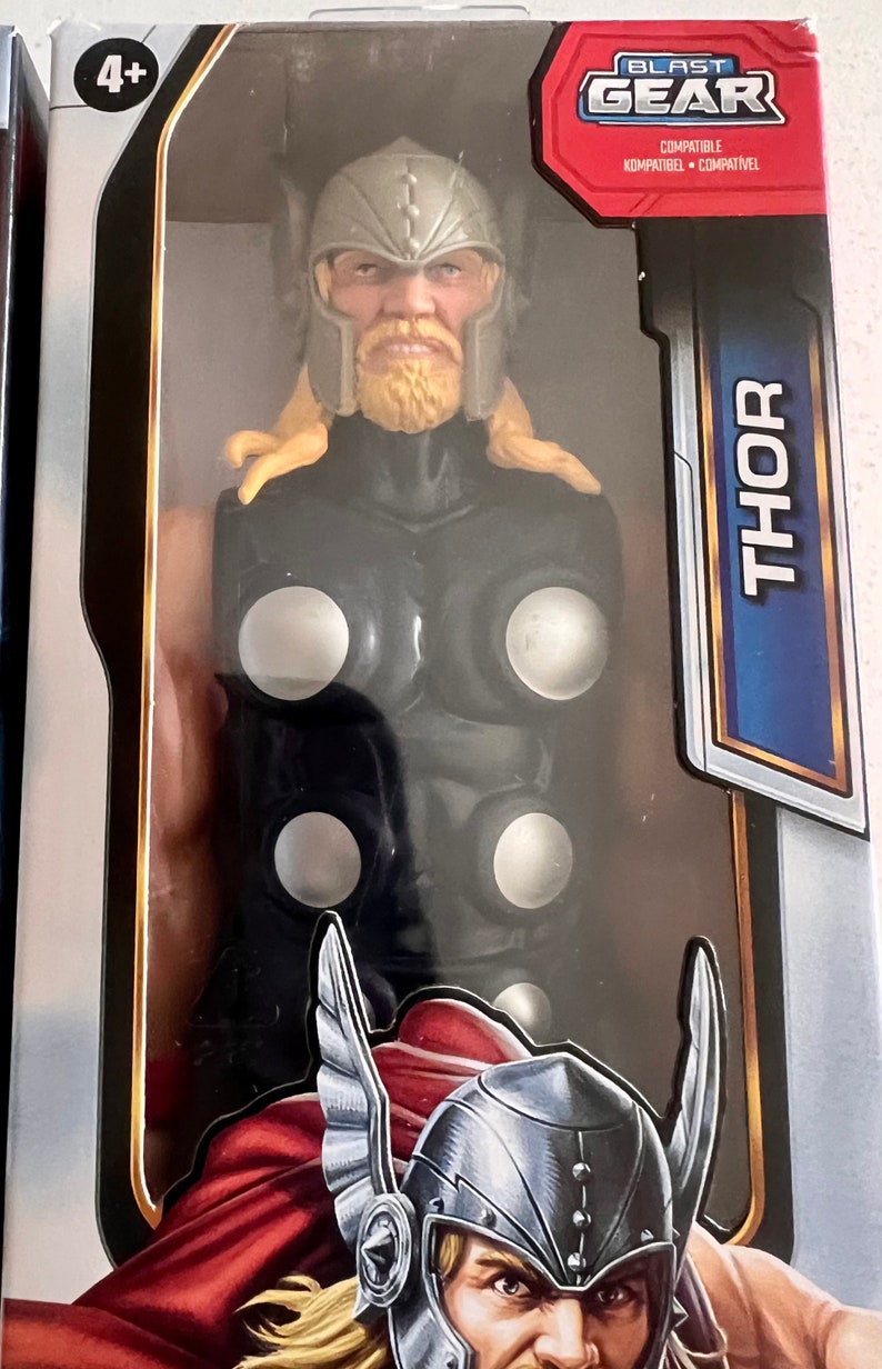 Pu&ograve; includere: Una action figure di Thor in una scatola. La figura ha un elmo argentato, capelli e barba biondi e un costume nero e argento. La scatola ha la scritta "Thor" in grandi lettere blu e il logo "Blast Gear".