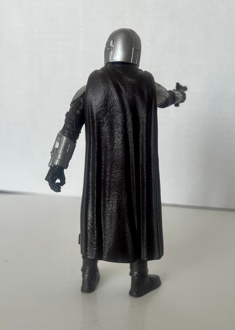 Puede incluir: Figura de acci&oacute;n con casco plateado y capa negra, sosteniendo una pistola bl&aacute;ster. La figura lleva una armadura y botas negras. La figura est&aacute; posando con el bl&aacute;ster apuntando hacia adelante.