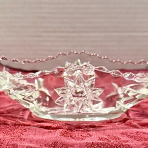 May include: Clear glass serving bowl with a starburst design in the centre. The bowl has a scalloped rim and a decorative, textured pattern. The bowl is on a red patterned surface.